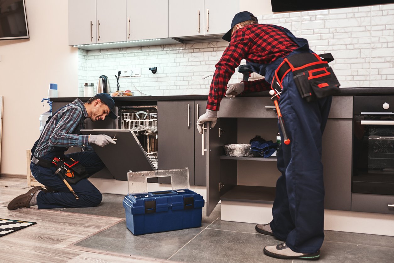 Learning from the best. Two men technician senior and young are sitting near dishwasher with screwdriver in kitchen with instruments Senior man teaches young one